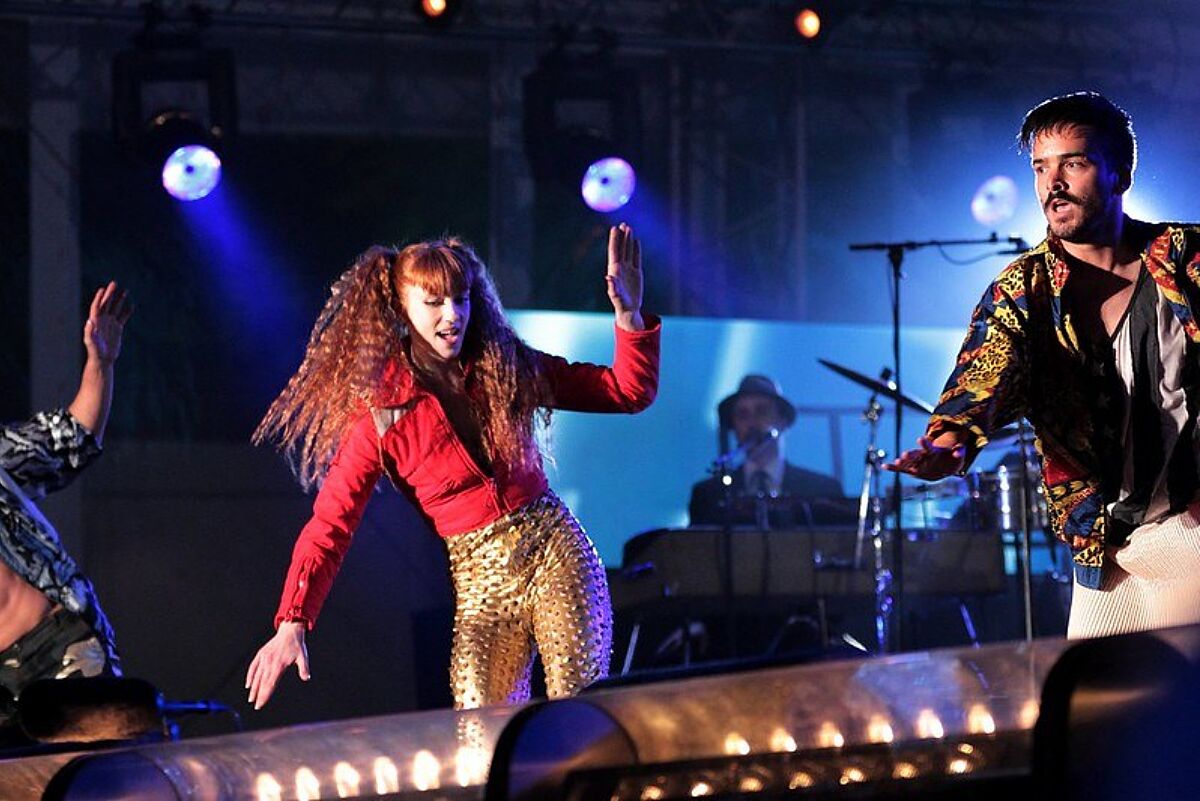 Livemuziek. Met een drummer, gitarist en dansende vrouw. Ze dragen felgekleurde kleding en staan op een podium waar meerder spotlights op gericht zijn.