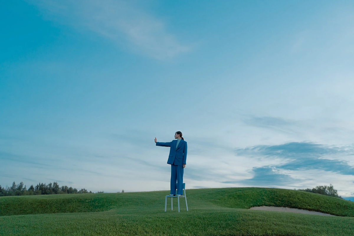 Person in a blue suit is standing in a green meadow and is signalling to stop. They're standing on top of a chair.