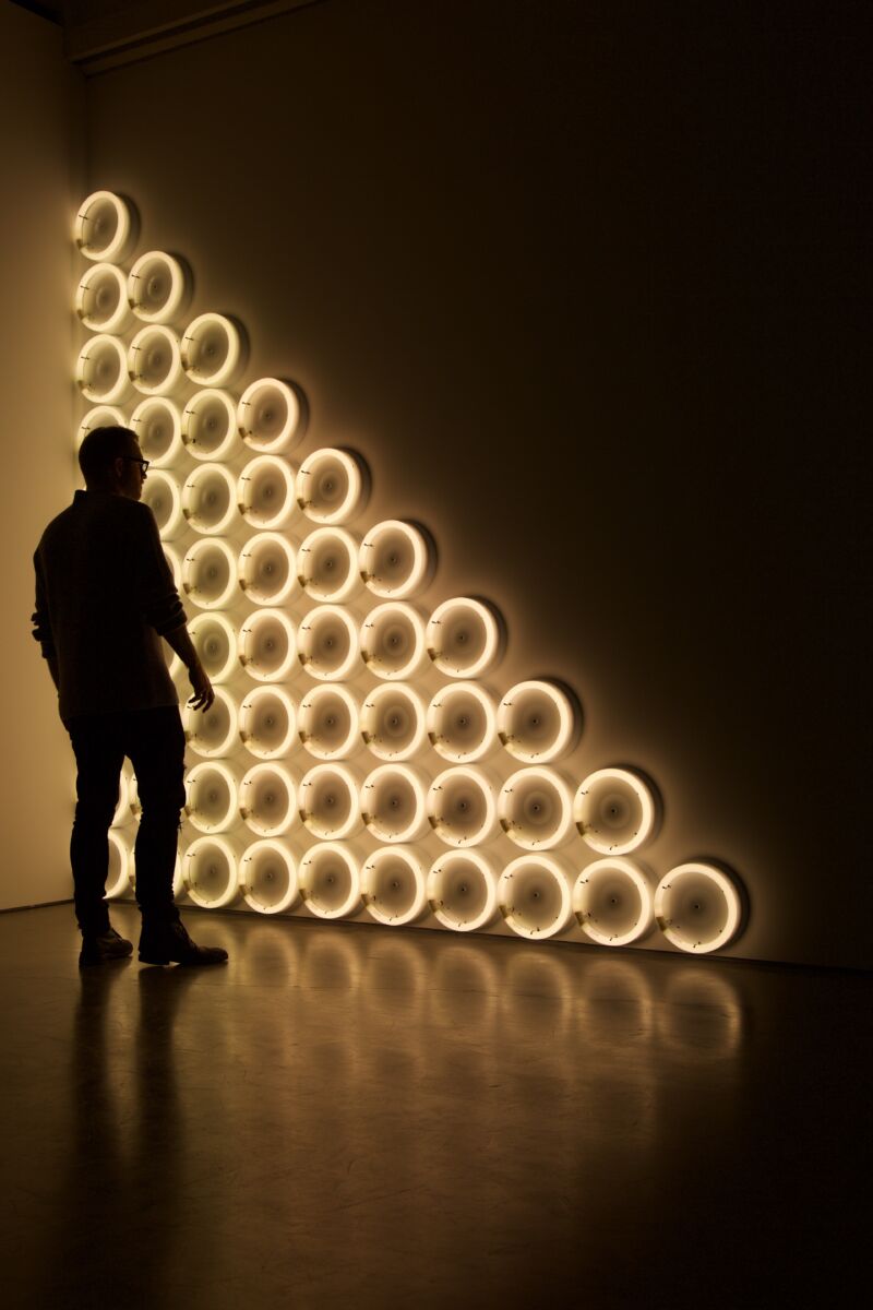 Persoon kijkt naar lichtgevende ringen die aan een muur zijn bevestigd in de vorm van een driehoek. Links tegen de wand zit de hoogste rij en elke rij rechts daarvan heeft één ring minder.